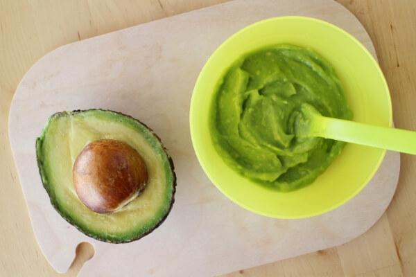 avocado half on a board with a bowl of mashed avocado and a baby spoon